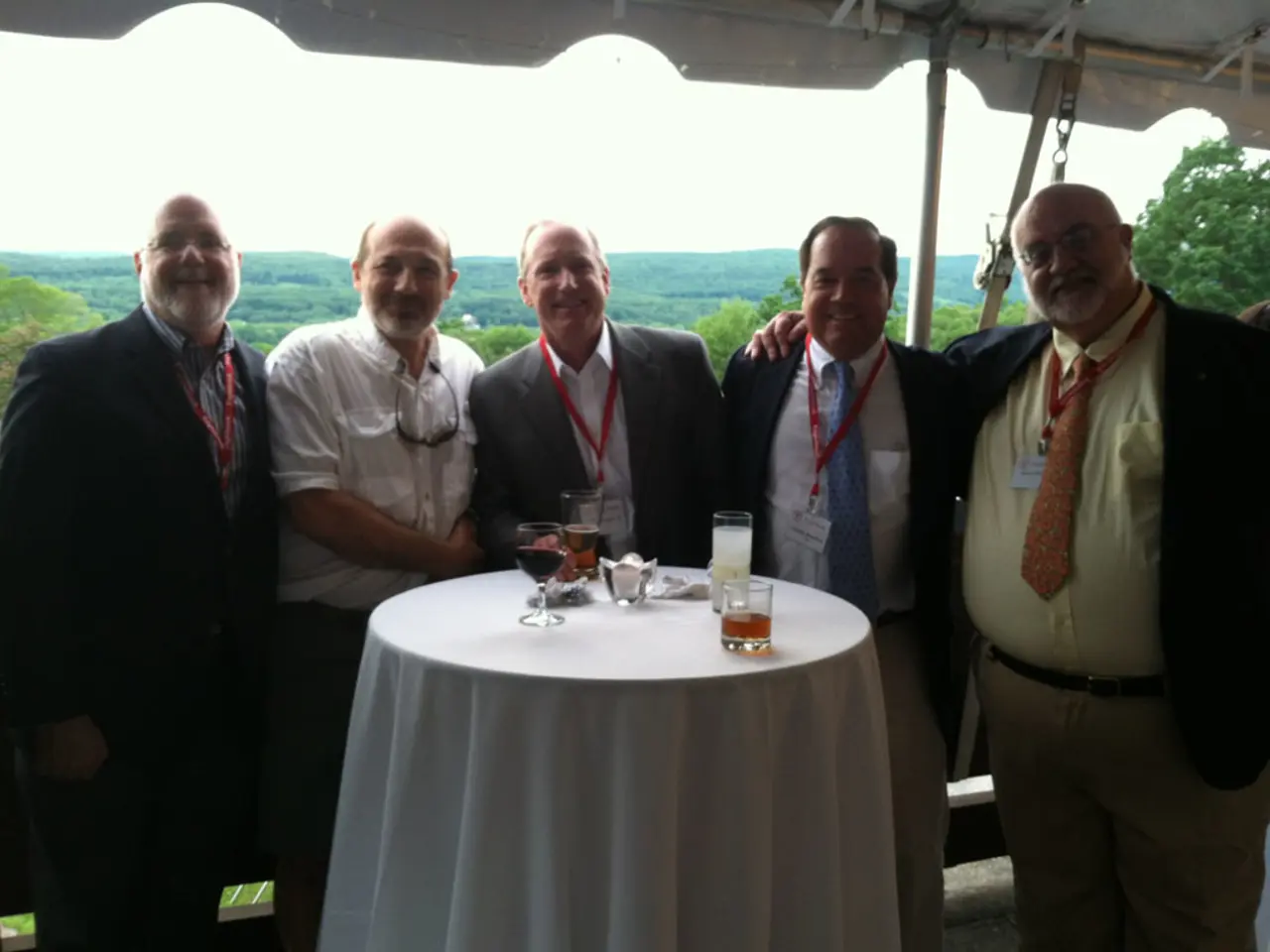 A group of five men are standing at a table with few glasses on it.