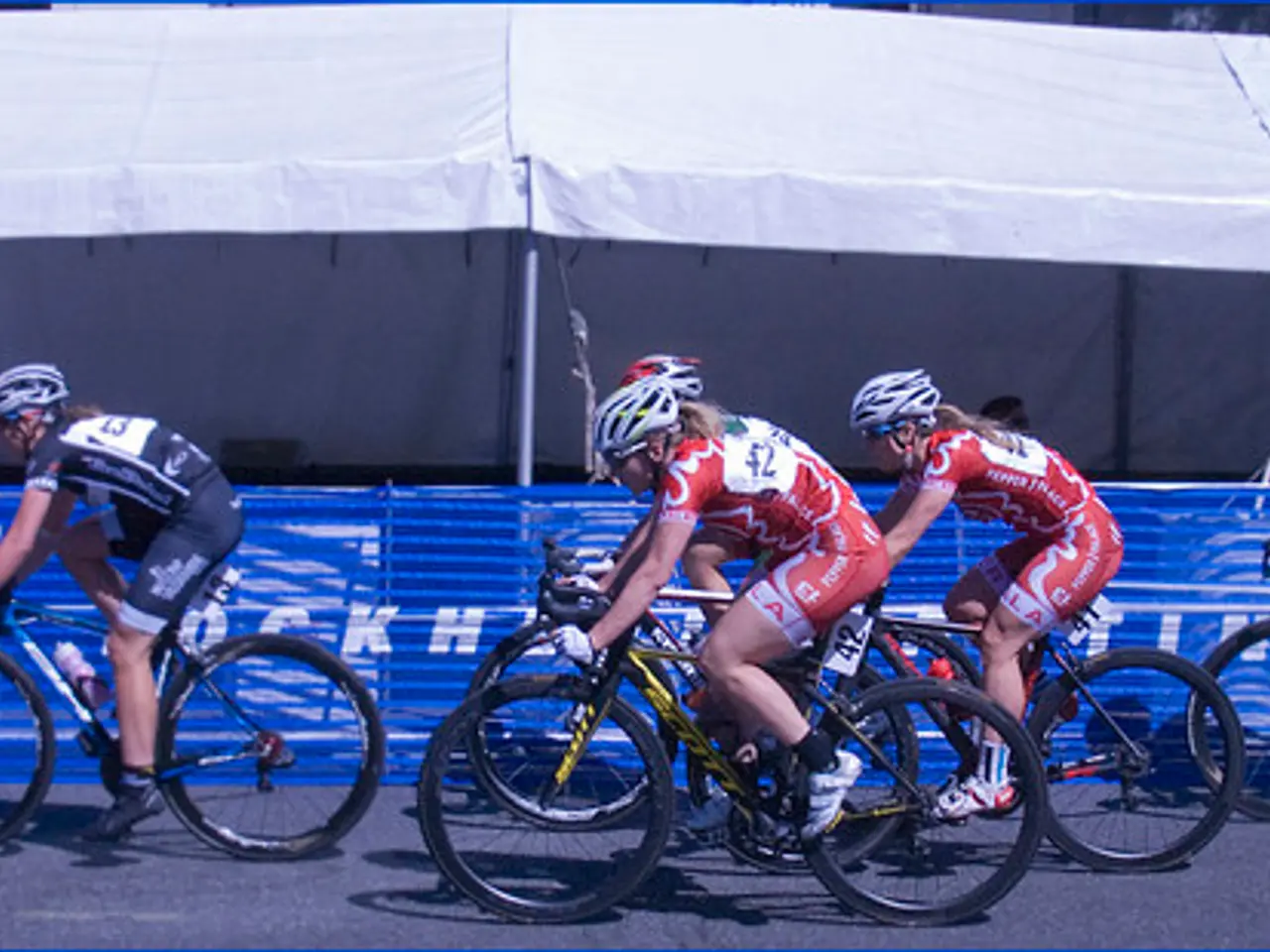 In the image few people are riding bicycles. Behind them there is fencing. Behind the fencing there...