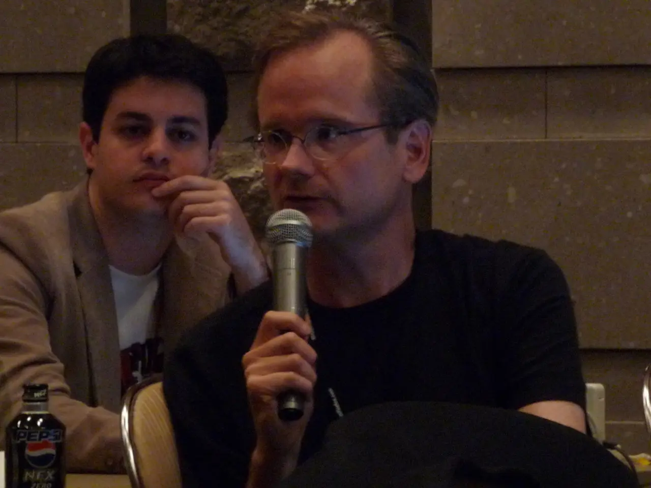 In this image I can see two people sitting on the chairs Among them one person is wearing the...