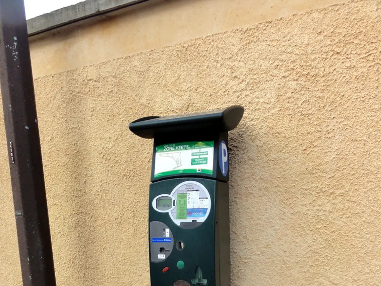 In the image there is an atm machine in front of the wall beside a pole.