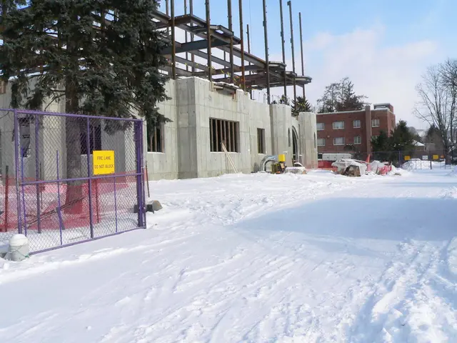 In this image land is covered with snow, in the background there are buildings, trees, fencing and...